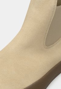 Bottine Chelsea en daim beige avec panneau latéral élastique et semelle en caoutchouc marron, montrée en gros plan sur fond blanc.