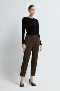 Vrouw in een zwart langslavend bovenstuk met geraffineerde details en bruine enkellange broek, met zwarte hakken, tegen een eenvoudige witte achtergrond.