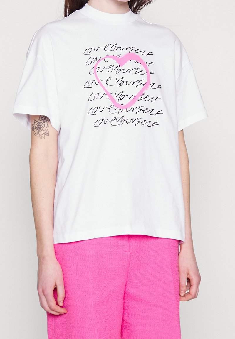 Hvid bomulds t-shirt med sort håndskrevet tekst "elsk dig selv" og et pink hjerte design. Korte ærmer og rund hals.