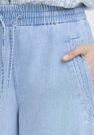 Hånd i lommen på lyseblå denim joggebukser med elastisk linning og snøre, båret med mønstret bluse.