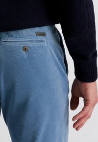 Close-up van een persoon wearing lichtblauwe corduroy broek met een achterzak met knoop en een mouw van een zwarte gebreide trui.