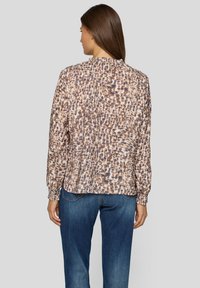 Braun-beige gemusterte Bluse mit langen Ärmeln, elastischen Bündchen und rundem Ausschnitt. Getragen zu blauen Jeans, die eine bequeme Passform zeigen.