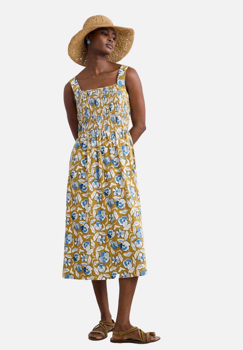 Bloemige midi-jurk met een gele achtergrond, blauwe en witte patronen, geraffineerde lijfje, vierkante halslijn en zakken. Gepaard met bruine sandalen met bandjes.