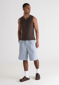 Brown ribbed tank top, light blue shorts, white socks, and brown clogs. Model stands with hands in pockets against a plain background.