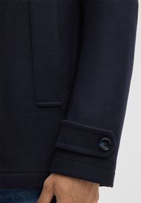 Dunkelblauer Mantelärmel mit einem geknöpften Riemen über dem Handgelenk, getragen über einer Hand und kombiniert mit blauen Jeans.