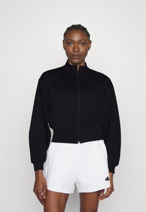 Giacca nera crop con zip, colletto alto e maniche lunghe, realizzata in tessuto morbido. Abbinata a pantaloni corti bianchi con un piccolo logo.