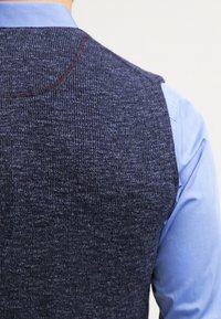 Mörkblå stickad väst med ribbad textur, med en hög krage och en slät blå skjorta under. Sömdetaljer är synliga.