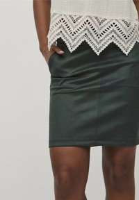 Žena v tmavě zelené sukně po kolena a bílé blůze s černou krajkovou lemovkou, ruka položená v kapse sukně.