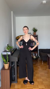 Γυναίκα με τατουάζ, φορώντας μαύρη ολόσωμη φόρμα χωρίς μανίκια και χνουδωτές παντόφλες, στέκεται σε ένα σαλόνι με πιάνο, φυτά, βιβλιοθήκη και καναπέ.