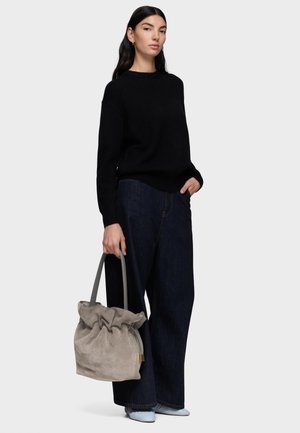 Donna con i capelli lunghi e scuri che indossa un maglione nero e jeans larghi scuri, che tiene in mano una borsa grigia morbida con due manici, in piedi su uno sfondo bianco.