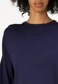 Maglione lavorato a maglia blu navy con scollatura rotonda e dettaglio del logo in rilievo sul davanti. Il tessuto appare morbido con una superficie liscia.