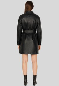 Donna con lunghi capelli castani, che indossa un cappotto in pelle nera con cintura e stivaletti neri, di spalle su uno sfondo grigio semplice.