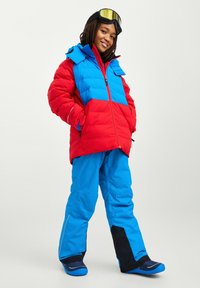 Tenue de ski hivernale colorée comprenant une doudoune rouge et bleue avec un col montant, associée à un pantalon de ski bleu et des chaussures de ski.