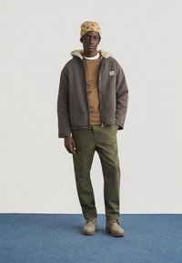 Άνδρας με καφέ μπουφάν, καφέ ζακέτα, χακί παντελόνι, μπεζ μποτάκια και κίτρινο σκουφάκι με καφέ βούλες που στέκεται σε μπλε χαλί.