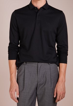 Homme portant un polo noir à manches longues avec des boutons et un pantalon gris habillé, debout les bras relâchés le long du corps.