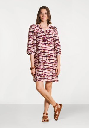 Femme portant une robe mi-longue à motifs rose et marron avec des manches trois-quarts, des sandales marron, un collier papillon et un bracelet marron, debout de manière détendue.