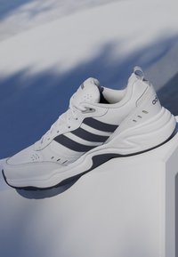 Baskets blanches Adidas à bandes noires présentées sur une surface blanche avec un fond flou bleu et blanc.