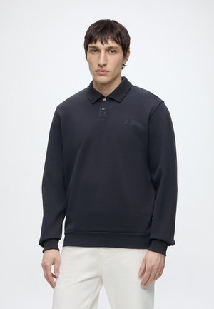 Jeune homme aux cheveux courts et foncés portant un polo noir à manches longues et un pantalon blanc, debout sur un fond blanc.