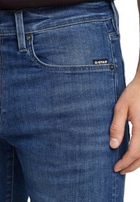Jeans din denim de culoare albastru închis G-Star, cu o croială slim, având un buzunar frontal, închidere cu buton metalic și o etichetă neagră cu marcă.