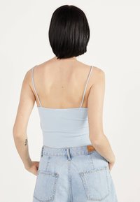 Mujer con pelo corto y negro, que lleva una blusa de tirantes de canalé de color azul claro y unos pantalones vaqueros de tiro alto de color azul claro con bolsillos.