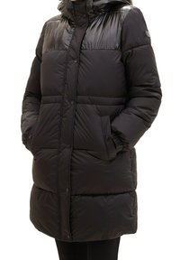 Czarna pikowana długa zimowa kurtka z zatrzaskami, kapturem i bocznymi kieszeniami, noszona przez osobę z rękami w kieszeniach.