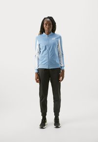 Svetlomodrá bunda na zips s bielymi rukávmi, čierne zúžené nohavice a čierne tenisky s zelenými akcentmi. Logo Puma viditeľné na oboch.