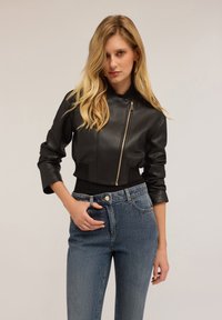 Czarna skórzana kurtka bomberka z zamkiem z przodu, krótkim krojem i ściągaczami na mankietach, w zestawieniu z niebieskimi dżinsami.