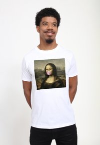 Muž v bílém tričku s potiskem Mona Lisy foukající růžovou bublinu z žvýkačky, stojící se založenýma rukama za zády, mírně se otočený do strany a usmívající se.