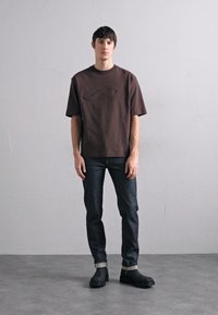 Camiseta de manga corta marrón con diseño texturizado, combinada con vaqueros de mezclilla oscuros y calzado negro con un acento gris en el puño.