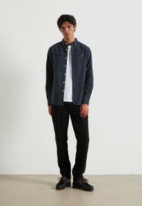Camicia a quadri blu navy e verde, sopra una maglietta bianca, abbinata a pantaloni neri e scarpe nere. Design semplice e vestibilità rilassata.