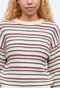 Personne portant un pull en tricot crème avec des rayures horizontales noires, roses et violettes, visible des épaules à la taille.