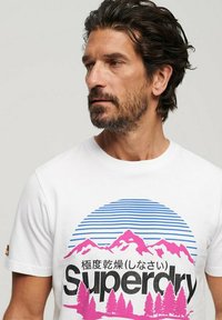 Camiseta de algodón blanca con un gráfico colorido de montañas en rosa y azul, con el texto "Superdry" y un fondo de rayas horizontales.