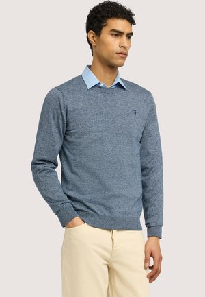 Junger Mann, der einen blauen Pullover mit Rundhalsausschnitt über einem hellblauen Hemd und beige Hose trägt, steht mit einer Hand in der Tasche.