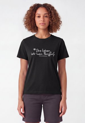 Jeune femme aux cheveux bouclés portant un t-shirt noir avec l'inscription "Das Leben ist kein Ponyhof" en écriture blanche manuscrite.
