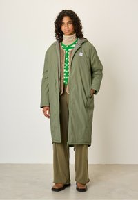 Długi, oliwkowozielony puchowy płaszcz z kapturem, na zamek i dwoma bocznymi kieszeniami. Nałożony na beżowy sweter dziewiarski z zielonymi kropkami.