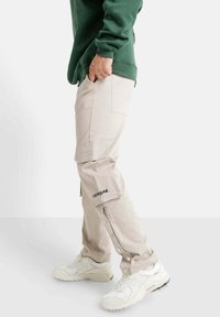 Beige cargobroek met knoopsluiting aan de zijkanten, gestikt logo-detail op het been, gecombineerd met witte sneakers en een groene sweatshirt.