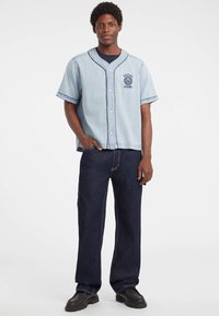 Lichtblauw denim overhemd met korte mouwen, donkere bies, knopen aan de voorkant en logo. Gepaard met donkerblauwe recht gesneden jeans en zwarte schoenen.