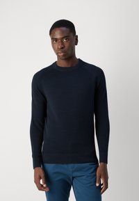 Marineblauer Strickpullover mit geripptem Rundhalsausschnitt und Bündchen, ausgestattet mit einem strukturierten Muster. Kombiniert mit blauen Hosen steht das Modell vor einem hellen Hintergrund.