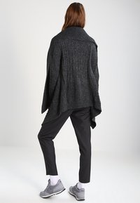 Tmavě šedý pletený pončo se širokým límcem a texturovaným vzorem, doplněný černými zúženými kalhotami a šedými sportovními botami.