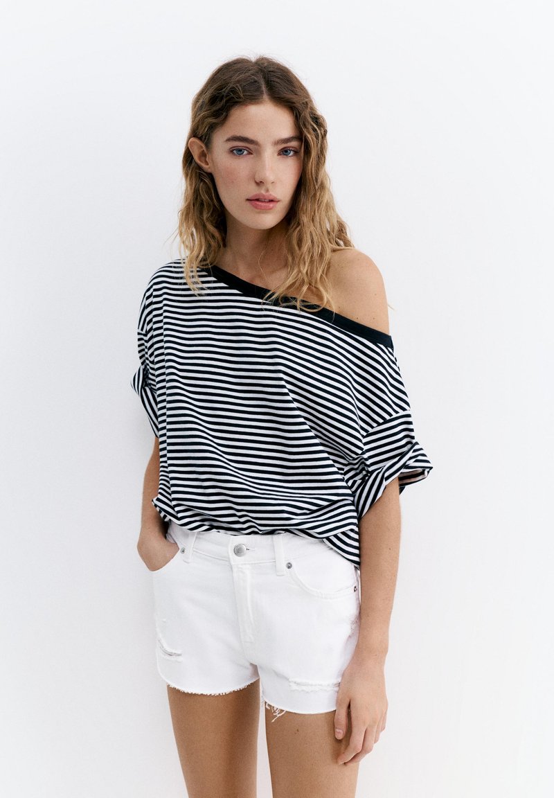 Jeune femme aux cheveux ondulés portant un haut rayé noir et blanc à épaules dénudées et un short blanc taille haute sur fond blanc.
