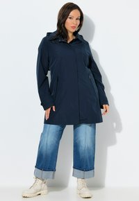 Mujer con impermeable azul marino con capucha, vaqueros azules de puños anchos y botas beige con cordones, de pie contra un fondo blanco.