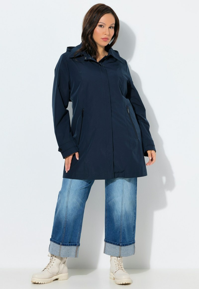 Mujer con impermeable azul marino con capucha, vaqueros azules de puños anchos y botas beige con cordones, de pie contra un fondo blanco.