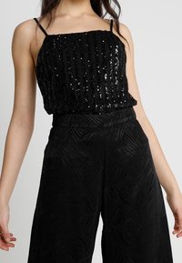 Svart paljett-topp med tunna band, med vertikala ränder, tillsammans med höga svarta byxor med ett strukturerat lövmönster.