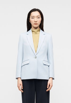 Kobieta w jasnoniebieskiej marynarce na jeden guzik, nałożonej na beżowy golf i czarne spodnie, stojąca na tle jednolitego jasnego tła.