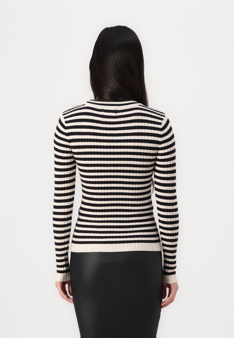 Femme aux longs cheveux noirs portant un pull ajusté à rayures horizontales noires et blanches et une jupe noire, debout de dos.