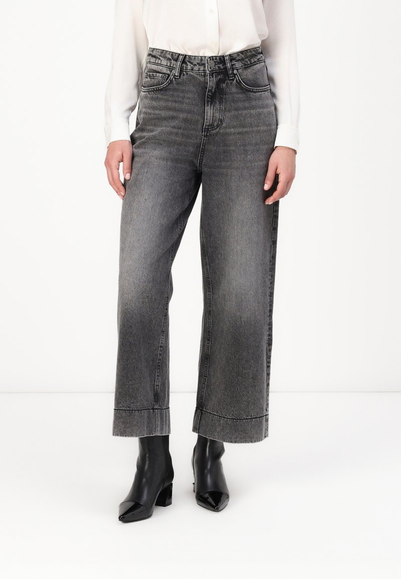 Femme portant un jean large noir délavé taille haute avec des ourlets retroussés, assorti à des bottines noires à bout pointu et un chemisier blanc.