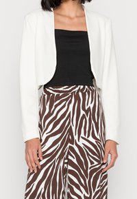 Valkoinen lyhyt takki, jossa on pitkät hihat, käytetty mustan yläosan päällä, yhdistetty ruskeisiin ja valkoisiin raitapolkka-housuihin.