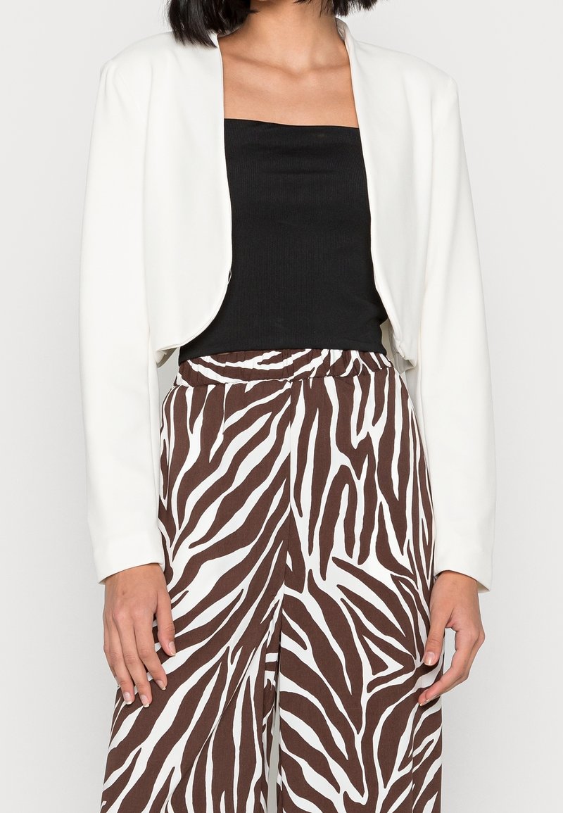 Valkoinen lyhyt takki, jossa on pitkät hihat, käytetty mustan yläosan päällä, yhdistetty ruskeisiin ja valkoisiin raitapolkka-housuihin.