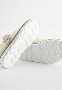 Kremowe buty o zaokrąglonym kształcie, z fakturowaną białą gumową podeszwą z wzorem w zygzak, oraz metalowym zapięciem po boku.