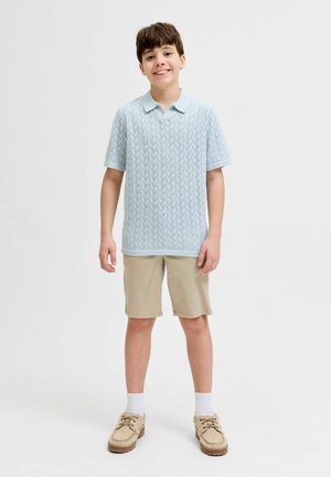 Garçon debout et souriant, portant un polo bleu clair texturé, un short beige, des chaussettes blanches et des mocassins beige.